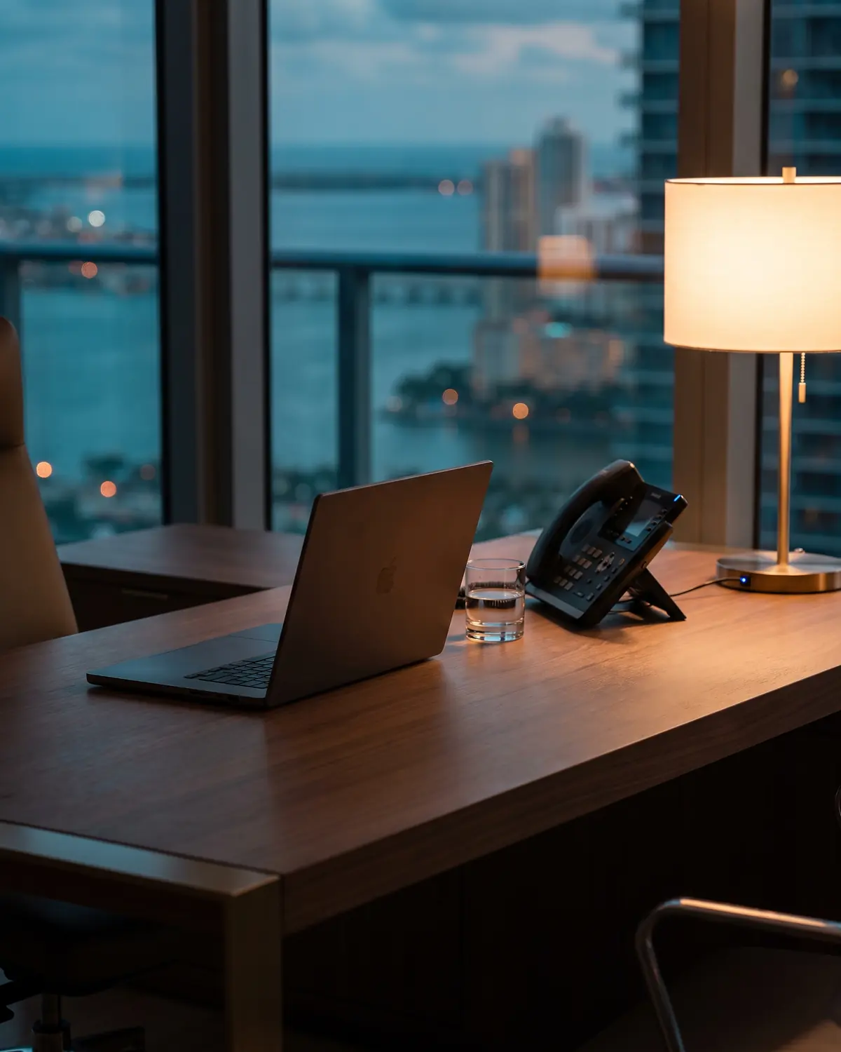 An executive desk with a laptop, an IP phone and a lamp in a high-rise office overlooking Miami at dusk.